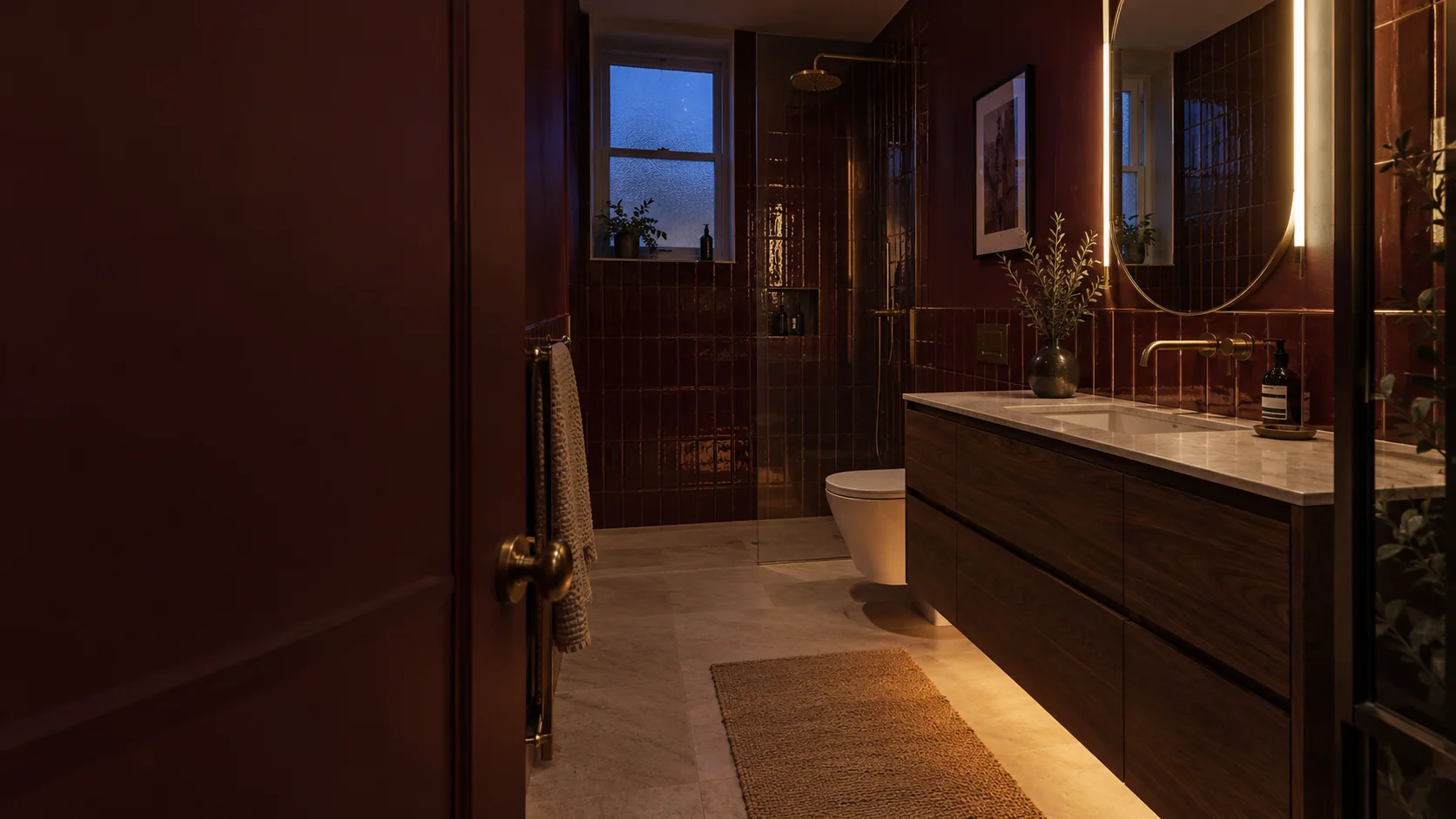 Warm mirror and low-level lighting in a burgundy bathroom with walnut cabinetry at dusk.