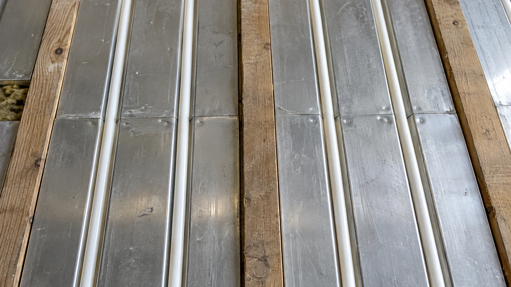 White heating pipe seated in aluminium spreader plates between timber floor joists.