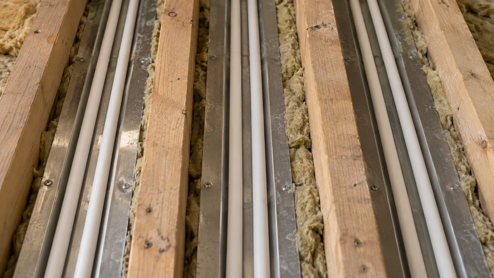 White heating pipes seated in aluminium spreader plates between timber joists above closely fitted insulation