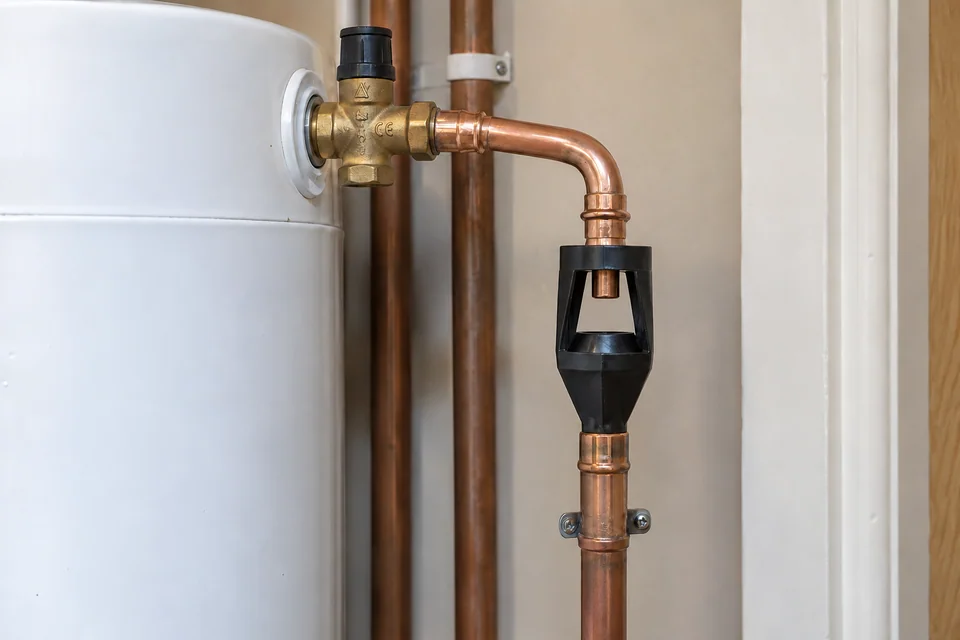 Close view of an open tundish and copper safety-discharge pipe beside a hot-water cylinder